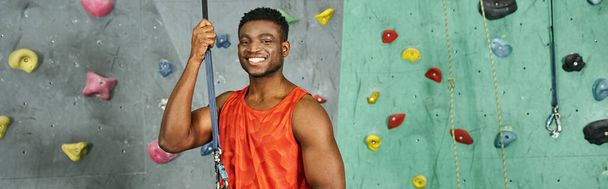 joyful athletic african american man in orange shirt smiling happily at camera, bouldering, banner - Photo, Image