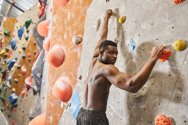 handsome shirtless african american man climbing up rock wall and looking at camera, bouldering - Photo, Image