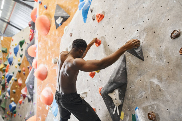 スポーティなトップレスのアフリカ系アメリカ人男性は,壁をボルダリングし,岩の上にグリップし,下を見下ろす - 写真・画像