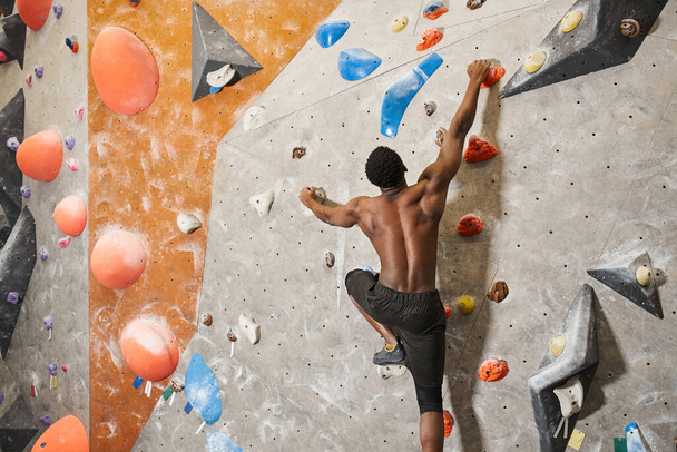 muscoloso uomo afroamericano senza maglietta che afferra fortemente i massi mentre si arrampica sulla parete rocciosa - Foto, immagini