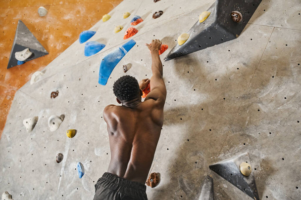sporty african american man with his shirt off reaching out boulder to grip, climbing up rock wall - Foto, Imagem