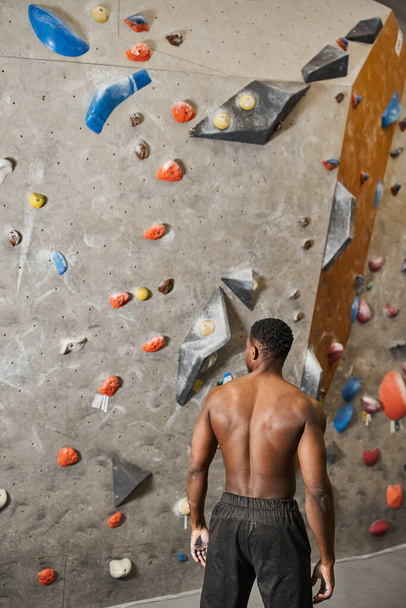 vertical shot of shirtless african american man in black pants posing next to bouldering wall - Photo, Image