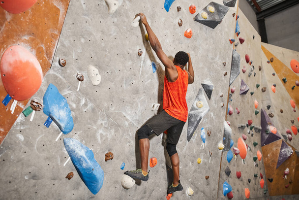 athletic african american man in orange shirt gripping on boulders while climbing up rock wall - Photo, Image
