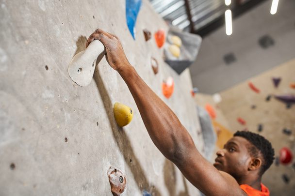 close up of handsome african american man gripping on boulders while climbing up rock wall - Photo, Image