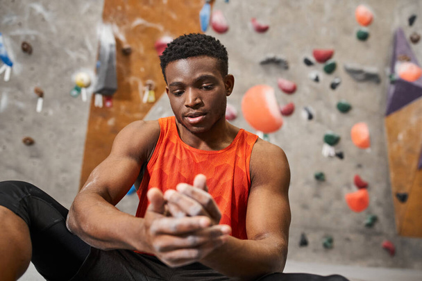 knappe Afro-Amerikaanse man in oranje shirt kijkt naar handen in gymnastiekkrijt met rotswand achtergrond - Foto, afbeelding