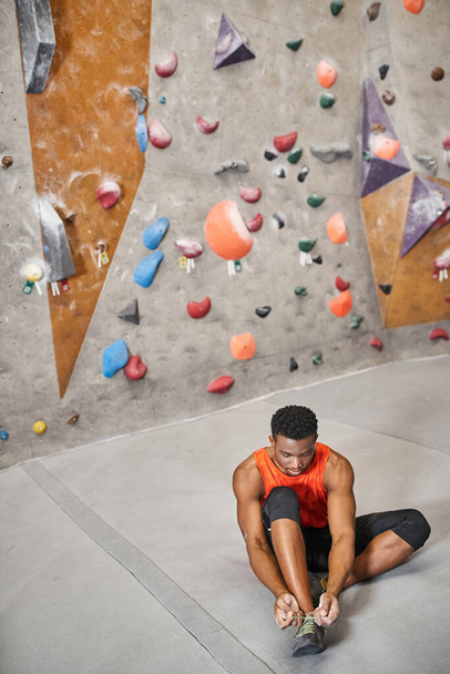 good looking african american man sitting on floor and tying his shoes next to climbing rock wall - Photo, Image