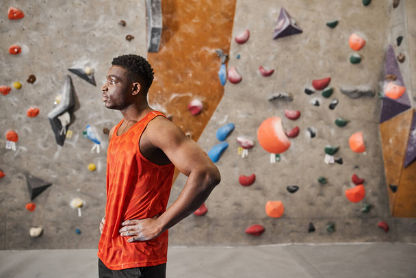 bonito afro-americano homem posando em perfil com as mãos em quadris ao lado de escalada parede de rocha - Foto, Imagem