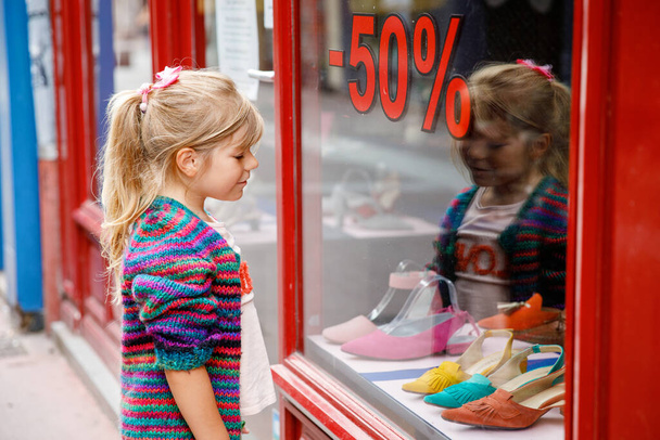 Маленькая девочка смотрит в окно возле торгового центра. Очаровательный счастливый ребенок смотрит на обувь в витрине магазина - Фото, изображение
