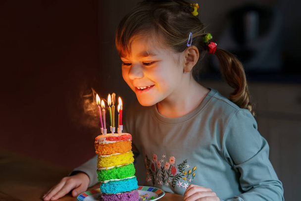 Счастливой маленькой дошкольницы, празднующей день рождения. Симпатичный улыбающийся ребенок с домашним радужным тортом, внутри. Счастливый здоровый малыш задувает шесть свечей на торте - Фото, изображение