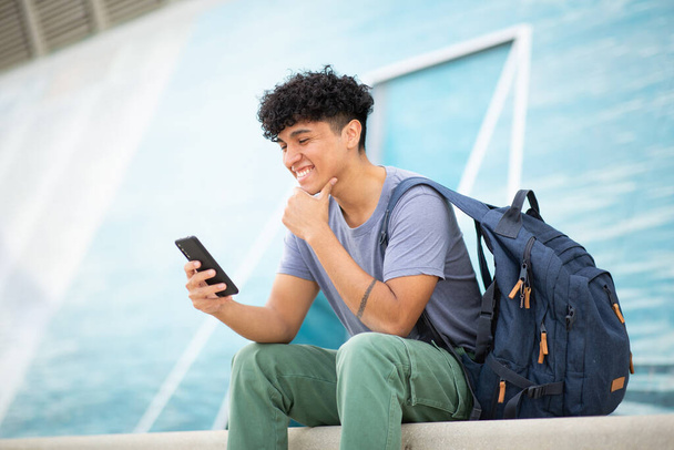 Портрет молодого улыбающегося человека, сидящего с мобильным телефоном и сумкой - Фото, изображение