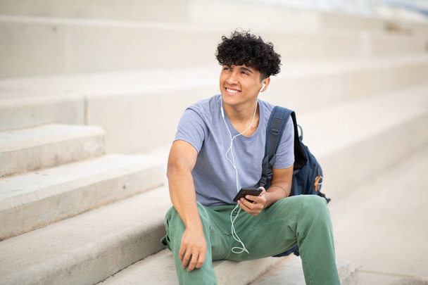 Портрет улыбающийся молодой человек, слушающий музыку с наушниками и мобильным телефоном - Фото, изображение