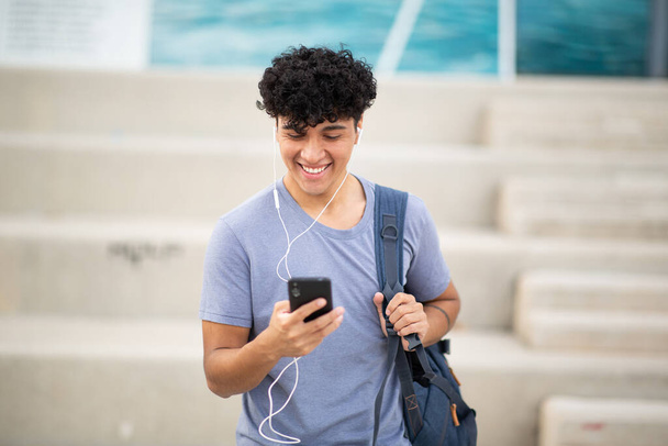Портрет улыбающийся молодой человек с наушниками, смотрящий на мобильный телефон - Фото, изображение