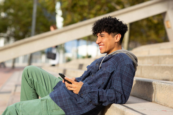 Боковой портрет улыбающийся молодой человек, сидящий на ступеньках на улице с сотовым телефоном - Фото, изображение