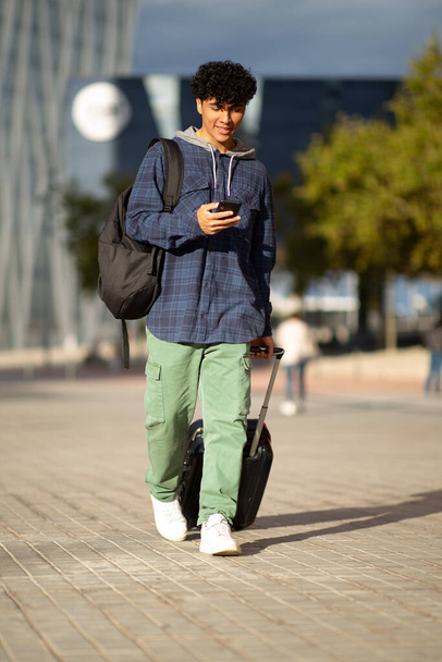 Ritratto completo del corpo giovane che cammina in città con cellulare e valigia - Foto, immagini