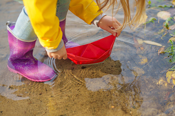 χαρούμενο κορίτσι με κίτρινο αδιάβροχο και χάρτινη βάρκα στο ποτάμι το φθινόπωρο στη φύση. - Φωτογραφία, εικόνα