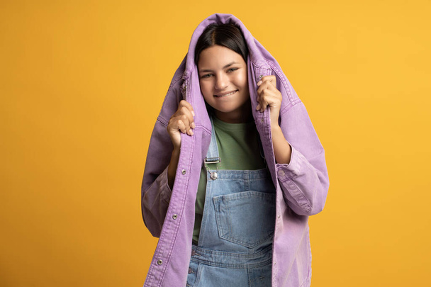Happy smiling playful teenager girl hiding with head in jeans purple jacket on yellow background. Friendly teen brunette girl wearing denim jumpsuit looking at camera. Advertisement banner, poster.  - Photo, Image
