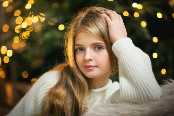 Piękna młoda dziewczyna z długimi kręconymi włosami stojąca na tle Bożego Narodzenia z lampkami boke i patrząc w dół. Magia ciepłe zdjęcie nowego roku. - Zdjęcie, obraz