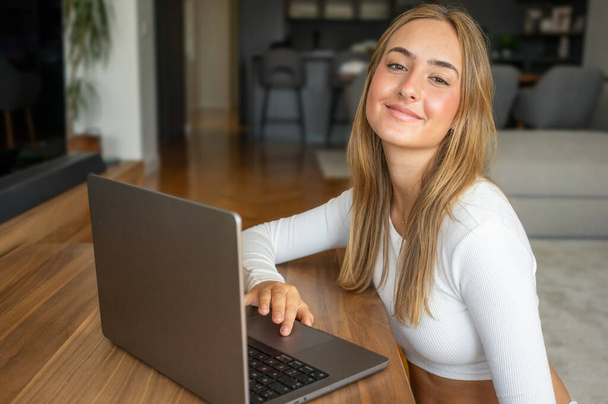 Lächelnde attraktive junge Frau, die auf dem Sofa sitzt und mit dem Laptop kommuniziert, zu Hause online arbeitet, fröhliches Teenie-Mädchen, das am Computer tippt, Blogs schreibt oder mit Freunden in sozialen Netzwerken chattet - Foto, Bild