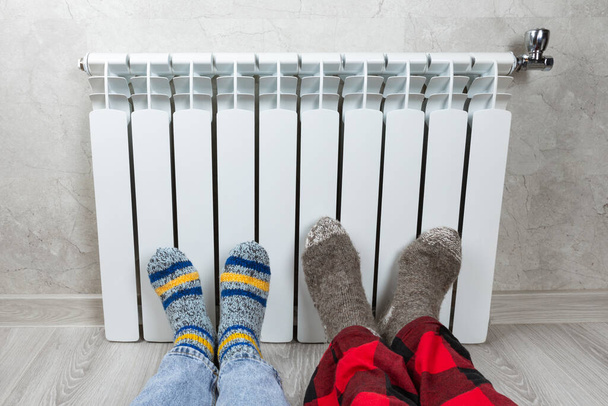 Pernas em um radiador contra o fundo de uma parede de luz. Homem e uma mulher em meias de malha quente de lã perto de um aquecedor doméstico na estação fria de outono-inverno. Conceito de estação de aquecimento em casa. - Foto, Imagem