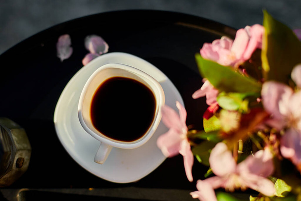 Вид зверху Чашка кавових квітів і блокнот на маленькому чорному столі на терасі вдома в сонячний день, відкритий робочий простір, літня релаксація, перерва на каву
. - Фото, зображення