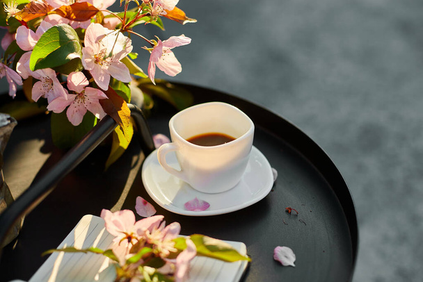 Kuppi kahvia kukkia ja muistikirja pieni musta pöytä terassilla kotona aurinkoisena päivänä, ulkona työtila, kesän rentoutumista, kahvitauko. - Valokuva, kuva