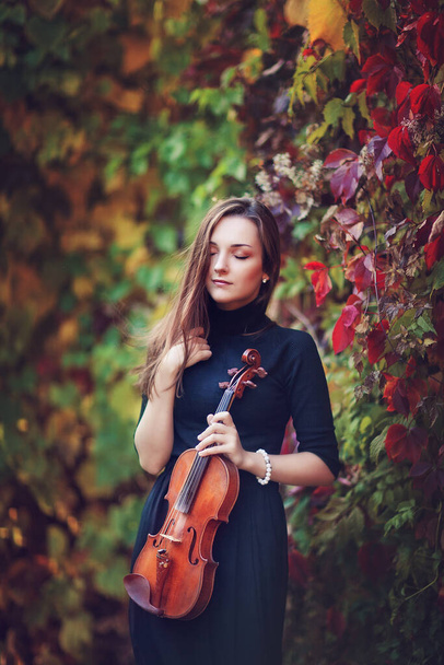 Portrait of a beautiful brunette girl in dark dress with violin. Violinist with close eyes in a autumn park. - Photo, Image
