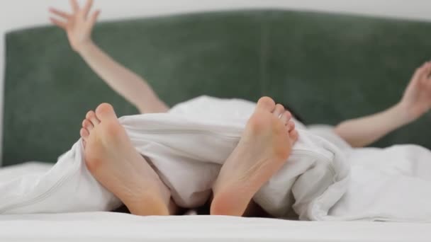 A nő lustán nyújtózkodik a királyméretű fehér ágyban. A női lábak a takaró alól látszanak. A pihenés és a nyaralás fogalma - Felvétel, videó