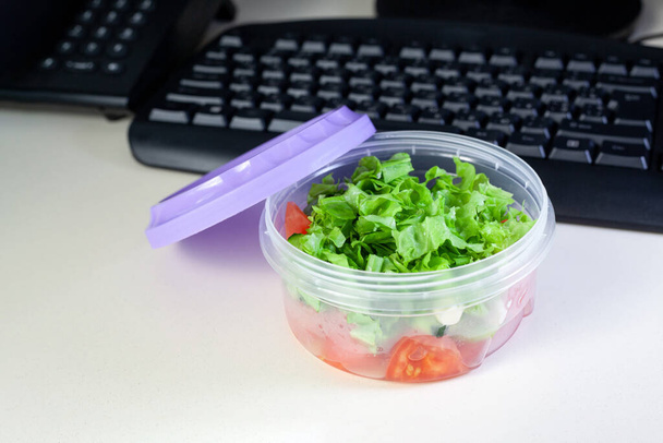 Γεύμα στο χώρο εργασίας. Δοχείο κουτί τροφίμων με φρέσκια σαλάτα στο γραφείο εργασίας με πληκτρολόγιο - Φωτογραφία, εικόνα