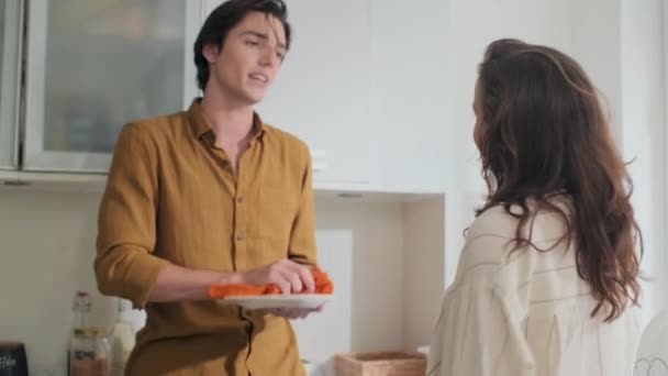 Молодой муж вытирает посуду, пока его жена разговаривает с ним и обнимает, стоя на кухне после ужина - Кадры, видео