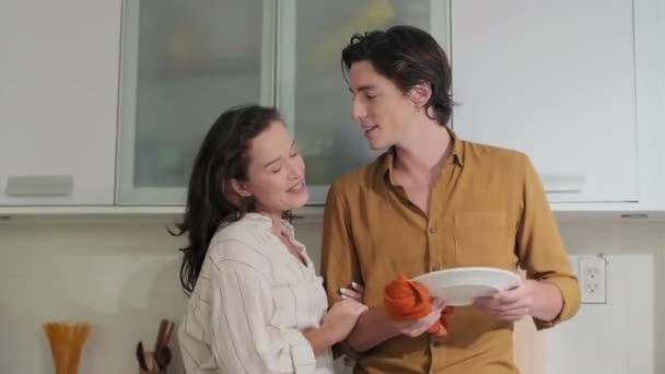 Közepes lövés fiatal boldog férfi megcsókolja a barátnője arcát, miközben letörli a tányért vacsora után a konyhában - Felvétel, videó