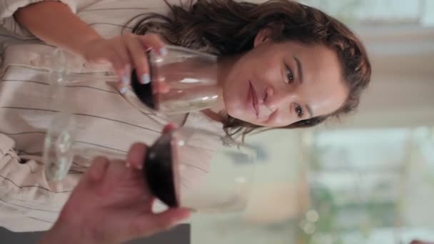 Tiro vertical de joven mujer alegre bebiendo vino tinto y tintineo vasos con su novio en la cena romántica en casa - Imágenes, Vídeo