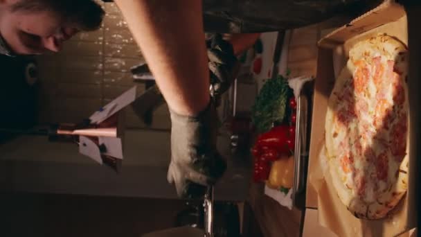 Colpo medio verticale di chef maschio caucasico tatuato che grattugia il parmigiano sopra la pizza appena sfornata in scatola di cartone da asporto, quindi la chiude - Filmati, video