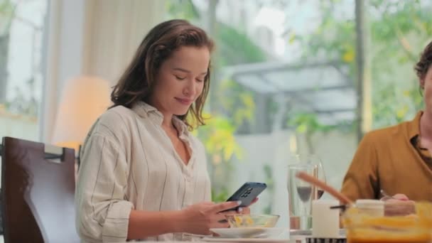Jovem mulher enviando mensagens de texto para alguém no celular enquanto se senta à mesa com o marido e janta em casa - Filmagem, Vídeo