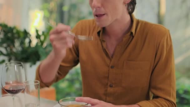 Tilt-up shot van Kaukasische man eten soep, wijn drinken en praten met iemand tijdens het eten thuis - Video