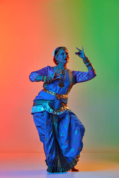 Odissi dance. Beautiful mature indian woman in traditional dress dancing indian dance against gradient studio background in neon light. Concept of beauty, fashion, India, traditions, choreography, art - Fotó, kép