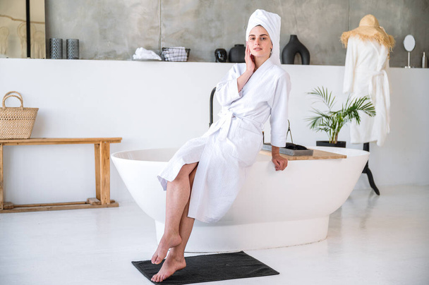 Beyaz bornozlu ve kafasında havlu olan genç bir kadın banyo küvetinin kenarında modern iç mimarisi ile oturuyor. Otel tuvaletinde bir kadın yüzüne dokunuyor ve duştan sonra dinleniyor. Vücut bakımı kavramı. - Fotoğraf, Görsel
