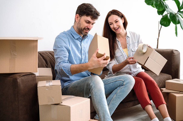 Happy young couple sitting on couch and packing household things in cartons. Using sticky tape. Prepare boxes to relocation to new house, flat or apartment. - Photo, Image