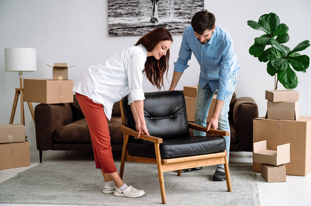 Glimlachend stel dat samen een moderne stoel draagt, meubels schikt, naar een nieuwe flat verhuist. Woninginrichting, inrichting woonkamer, renovatie en renovatie - Foto, afbeelding