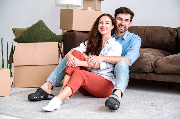 Koncept přemísťování. Šťastný mladý muž a žena se stěhují v novém nájemním domě spolu, vybalují krabice a odpočívají na podlaze vedle pohovky v obývacím pokoji - Fotografie, Obrázek