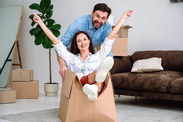Happy young man pushing cardboard box with smiling woman in new living room. Excited married couple having fun, celebrating moving into apartment. - Photo, Image