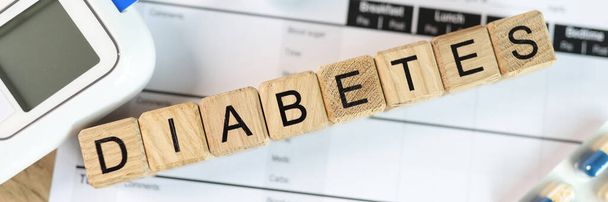 Word diabetes is assembled from wooden cubes with pills, glucometer and blood sugar tests on medical form paper. Concept of diabetes and blood sugar self-control. - 写真・画像