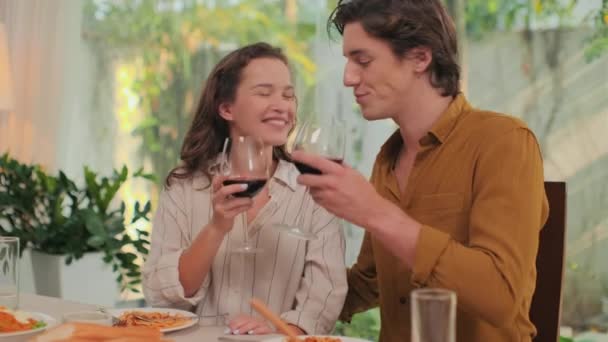 Średnie ujęcie młodej pary pijącej czerwone wino ze szklanek i całującej się podczas romantycznej kolacji w domu - Materiał filmowy, wideo