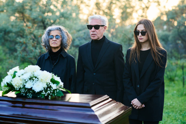 Familia rota de tres en ropa de luto y gafas de sol de pie frente al ataúd cerrado con su pariente muerto o buen amigo - Foto, Imagen