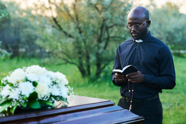 Afro-Amerikaanse pastoor met open bijbel het uitvoeren van begrafenisdienst op kerkhof terwijl staan bij houten kist met witte bloemen op de top - Foto, afbeelding