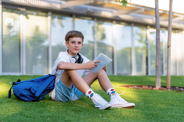Teini-ikäinen poika katselee kameraa istuessaan nurmikolla muistikirjan ja repun kanssa. Koulutuksen käsite. - Valokuva, kuva