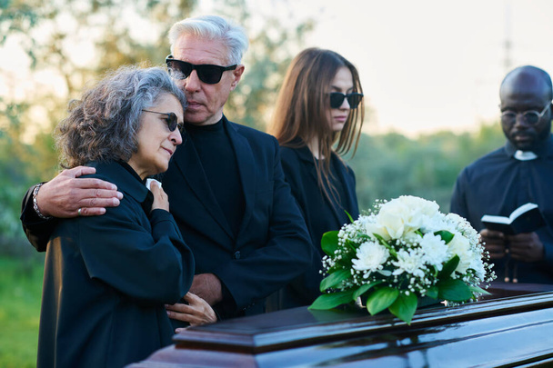 Familia de luto de tres y sacerdote en traje negro de pie frente al ataúd con flores blancas en la parte superior durante el funeral en el cementerio - Foto, Imagen