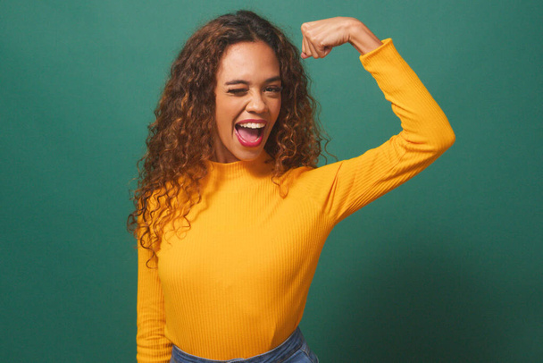Potente mujer fuerte flexiona el músculo del brazo, guiña sonrisas, verdes fondo del estudio. Foto de alta calidad - Foto, Imagen