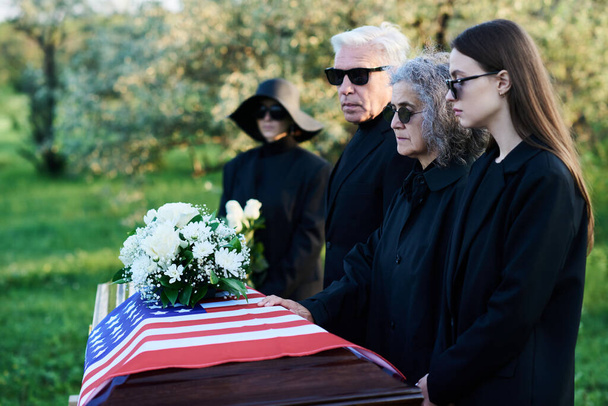 Güneş gözlüklü ve yas tutan aile fertleri Amerikan bayrağıyla kaplanmış tabutun başında durup ölen akrabalarının yasını tutuyorlar. - Fotoğraf, Görsel