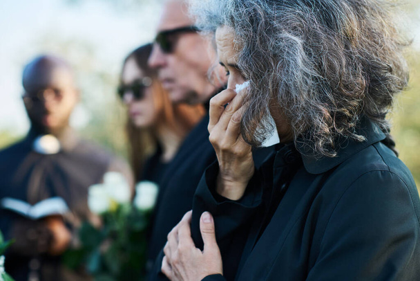 Mujer mayor con el pelo gris limpiando lágrimas con el pañuelo en el funeral de su pariente o miembro de la familia mientras está de pie delante de la cámara - Foto, Imagen