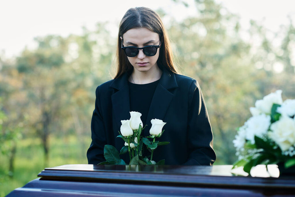 Young widow with bunch of fresh white roses standing by coffin with closed lid and flowers on its top while grieving about her dead husband - Photo, Image